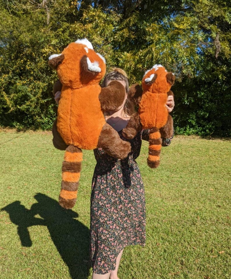 May include: A person holding two plush red panda toys. The larger toy is brown and orange with a white face and black stripes on the tail. The smaller toy is a similar colour and design but is smaller in size.