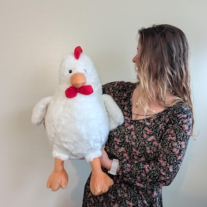 May include: A large plush chicken toy with white fur, a bright red comb on top of its head, a red wattle under its orange beak, and two orange feet. The stuffed animal has small black eyes and a rounded body, featuring soft, textured white fabric.