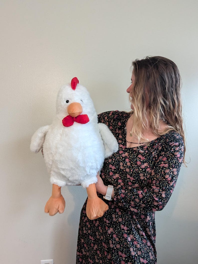 May include: A large plush chicken toy with white fur, a bright red comb on top of its head, a red wattle under its orange beak, and two orange feet. The stuffed animal has small black eyes and a rounded body, featuring soft, textured white fabric.