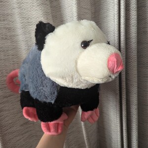May include: A plush toy of a possum with black and gray fur, a white face, and pink nose and feet.