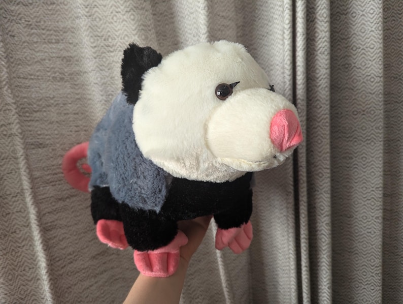 May include: A plush toy of a possum with black and gray fur, a white face, and pink nose and feet.