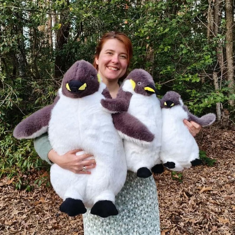 May include: Three plush penguin toys of varying sizes, each featuring a dark gray back, white belly, black feet, and a yellow patch near the beak. The penguins have soft, fuzzy textures and are arranged from largest to smallest in a row.