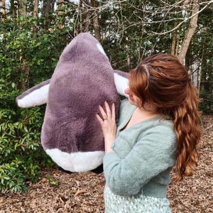 May include: A large, plush penguin toy featuring soft, dark gray fur on its back and head, with contrasting white fur on its belly and wing tips. The stuffed animal has a rounded, plump body shape and is held upright, showcasing its textured fabric.