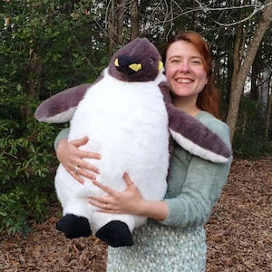 May include: A large plush penguin toy featuring a soft, dark gray head and back, a white belly, and black feet. The penguin has a small beak with yellow accents near the base. The plush is constructed with a fuzzy, textured fabric throughout.