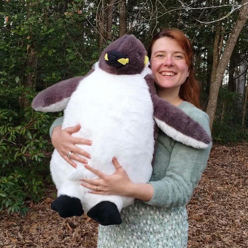 May include: A large plush penguin toy featuring a soft, dark gray head and back, a white belly, and black feet. The penguin has a small beak with yellow accents near the base. The plush is constructed with a fuzzy, textured fabric throughout.