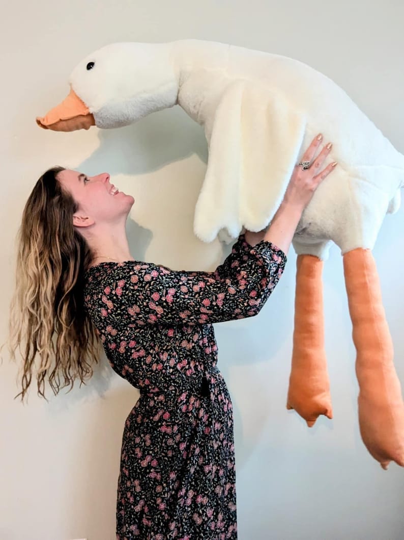 K&ouml;nnte beinhalten: Eine Frau in einem schwarzen Blumenkleid h&auml;lt eine gro&szlig;e wei&szlig;e Pl&uuml;schgans mit orangenen Beinen. Die Gans schaut die Frau an.