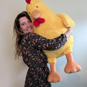 May include: A large, plush yellow chicken stuffed animal with a soft, fuzzy texture. The toy features a bright red comb on top of its head, a red wattle under its beak, a tan-colored beak, and two large, flat, orange feet dangling downwards.