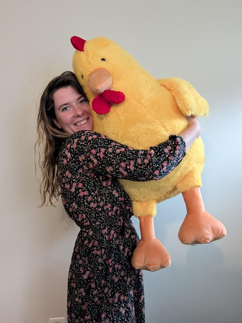 May include: A large, plush yellow chicken stuffed animal with a soft, fuzzy texture. The toy features a bright red comb on top of its head, a red wattle under its beak, a tan-colored beak, and two large, flat, orange feet dangling downwards.