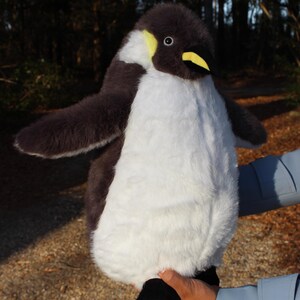 May include: A plush penguin toy featuring soft dark gray and white fur, a yellow patch near the eye, and a yellow beak. The stuffed animal has outstretched wings and black feet, designed with a realistic appearance and a rounded, plump body shape.