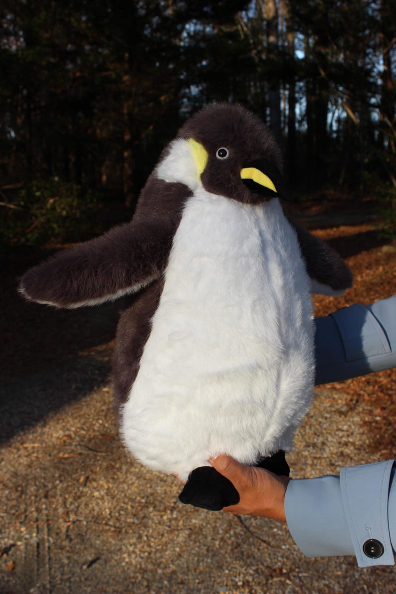 May include: A plush penguin toy featuring soft dark gray and white fur, a yellow patch near the eye, and a yellow beak. The stuffed animal has outstretched wings and black feet, designed with a realistic appearance and a rounded, plump body shape.