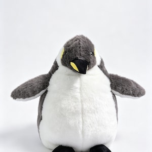 May include: A plush penguin toy featuring soft dark gray fur on the head, back, and wings, with a contrasting white belly. The penguin has a black beak with a yellow accent near the base and small black feet, standing in an upright position.