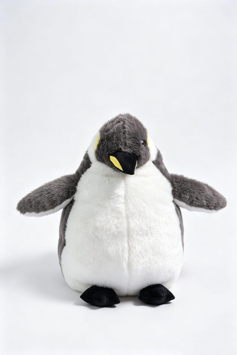May include: A plush penguin toy featuring soft dark gray fur on the head, back, and wings, with a contrasting white belly. The penguin has a black beak with a yellow accent near the base and small black feet, standing in an upright position.