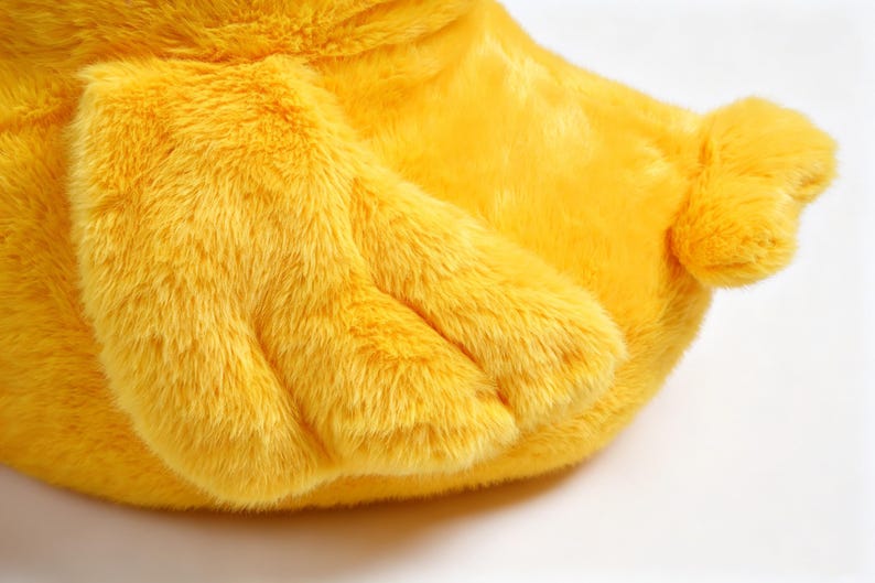 May include: A bright yellow plush toy featuring soft, synthetic fur texture. The object displays a prominent wing-like appendage with three distinct segments and a small, rounded tail section extending from the main body of the stuffed animal.