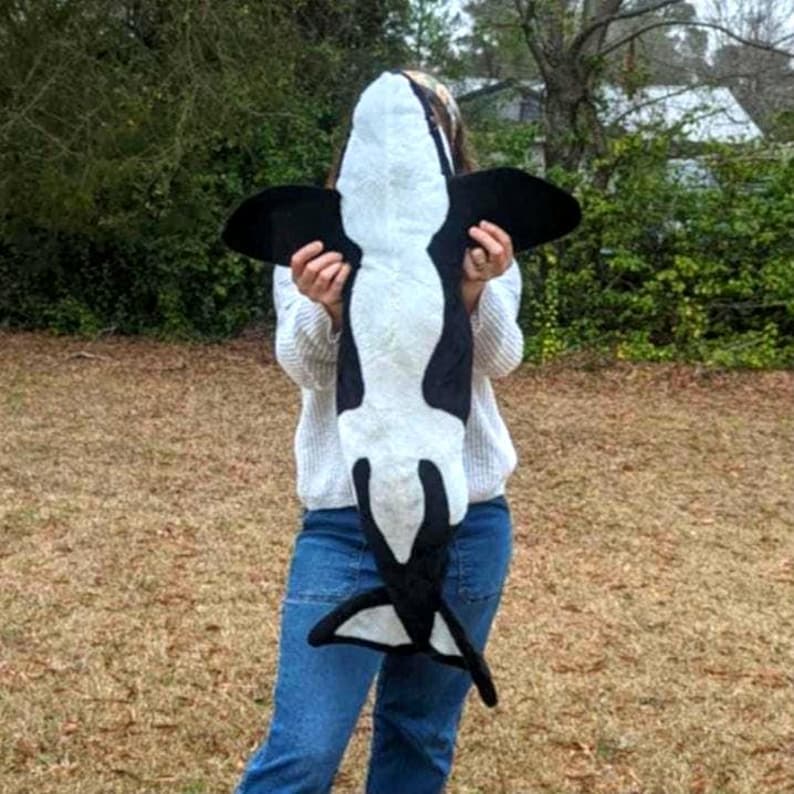 May include: A plush, black and white orca whale stuffed animal. The toy has a white belly and a black back, with black fins and tail. The orca is held up in front of a person.