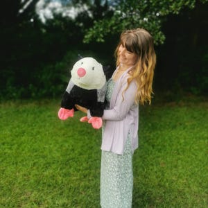 May include: A woman in a light purple cardigan and a green floral dress holds a large plush opossum with black and white fur and pink feet.