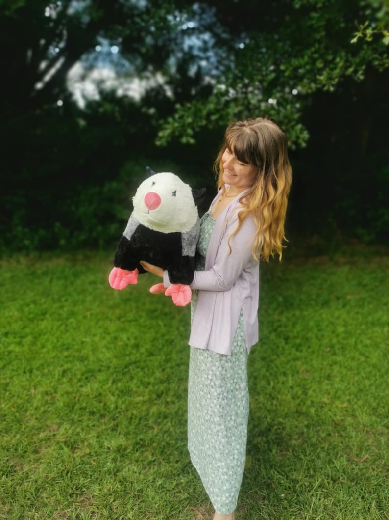 May include: A woman in a light purple cardigan and a green floral dress holds a large plush opossum with black and white fur and pink feet.