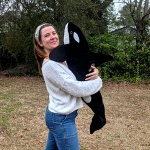 May include: A plush orca whale toy in black and white is held in front of a person. The stuffed animal has white patches and a smiling expression. The background is a blurred outdoor scene with green foliage.