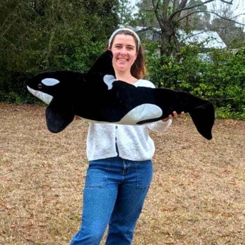 May include: A plush orca whale toy, primarily black with white markings on its belly and eyes. The toy is soft and cuddly, designed to resemble a killer whale. The orca toy is a popular item for children and adults alike.