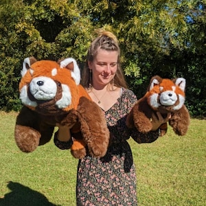 May include: A woman is holding two plush red panda toys. The larger toy is brown and orange with a white face, and the smaller toy is a similar colour but smaller in size.