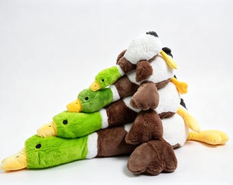 May include: A stack of four plush mallard duck toys arranged in a pyramid shape. Each duck features a bright green head, a yellow beak, a white neck ring, and a brown body with soft, fuzzy texture. The ducks are stacked from largest to smallest.