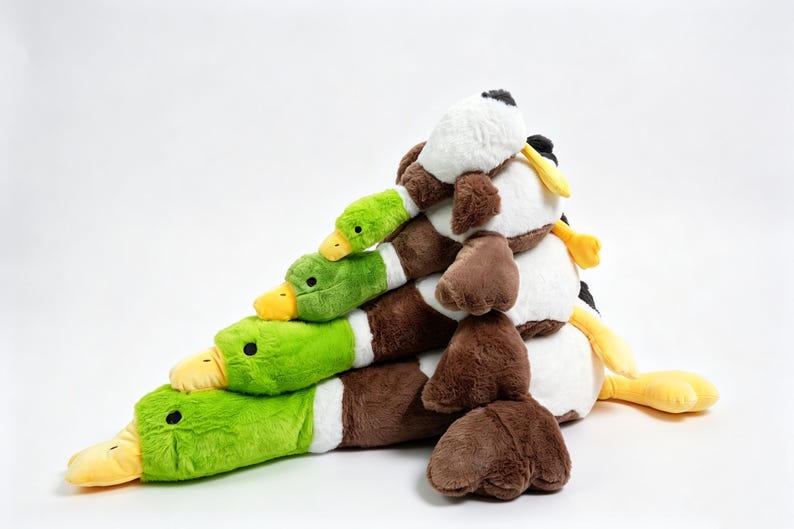 Puede incluir: Una pila de cuatro juguetes de peluche con forma de pato real dispuestos en forma de pir&aacute;mide. Cada pato presenta una cabeza de color verde brillante, un pico amarillo, un anillo blanco en el cuello y un cuerpo marr&oacute;n con textura suave.