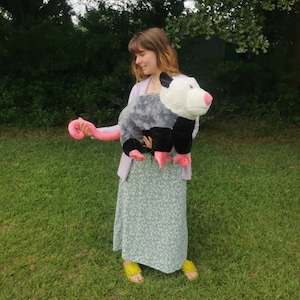 May include: A person holding a large plush opossum toy. The opossum is gray and black with a pink tail and white face. The person is wearing a light blue dress with a floral pattern and yellow sandals.
