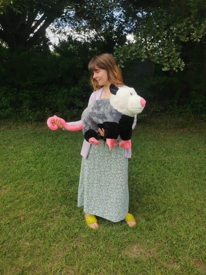 May include: A person holding a large plush opossum toy. The opossum is gray and black with a pink tail and white face. The person is wearing a light blue dress with a floral pattern and yellow sandals.