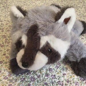 K&ouml;nnte beinhalten: Ein Pl&uuml;sch-Waschb&auml;r-Spielzeug mit grauem, wei&szlig;em und braunem Fell. Das Spielzeug hat ein detailliertes Gesicht mit schwarzen Augen und einer schwarzen Nase. Der Waschb&auml;r liegt auf einer Oberfl&auml;che mit floralem Muster.