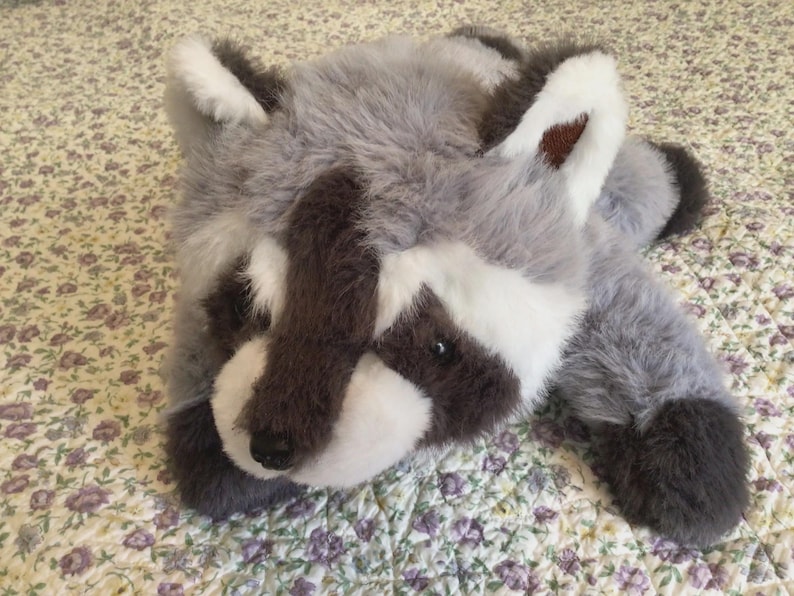 K&ouml;nnte beinhalten: Ein Pl&uuml;sch-Waschb&auml;r-Spielzeug mit grauem, wei&szlig;em und braunem Fell. Das Spielzeug hat ein detailliertes Gesicht mit schwarzen Augen und einer schwarzen Nase. Der Waschb&auml;r liegt auf einer Oberfl&auml;che mit floralem Muster.