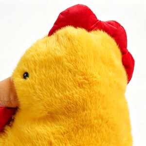 May include: A plush chicken toy featuring bright yellow fur, a vibrant red comb on top of the head, a red wattle under the beak, and a small brown beak. The toy has a soft, fuzzy texture and a round, stuffed shape, captured from a side profile.