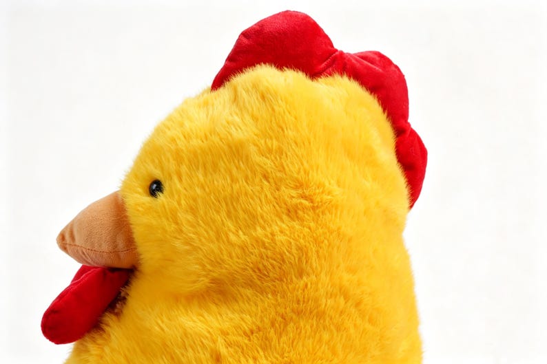 May include: A plush chicken toy featuring bright yellow fur, a vibrant red comb on top of the head, a red wattle under the beak, and a small brown beak. The toy has a soft, fuzzy texture and a round, stuffed shape, captured from a side profile.