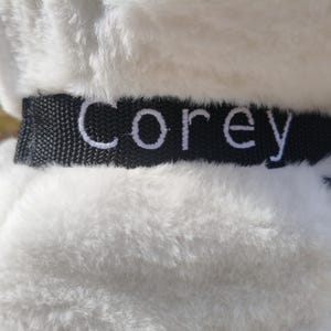 May include: Close-up of a white plush toy with a black collar. The word "Corey" is embroidered in white on the collar. A black zipper is visible on the side of the toy.