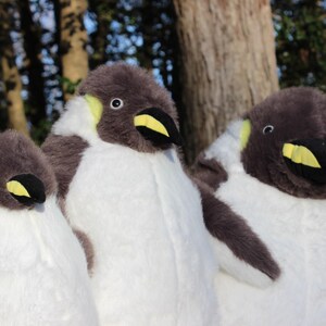 May include: Three plush penguin toys featuring soft grey and white fur, black beaks with yellow accents, and small round eyes. The stuffed animals are arranged side by side, showcasing their rounded bodies and detailed facial features in a set.