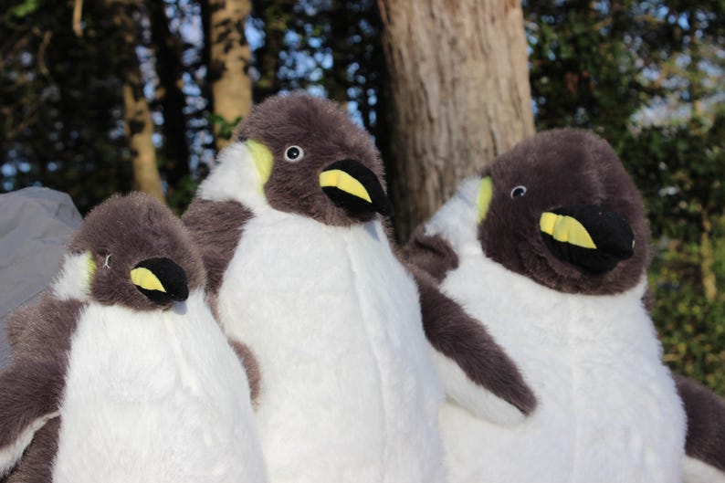 May include: Three plush penguin toys featuring soft grey and white fur, black beaks with yellow accents, and small round eyes. The stuffed animals are arranged side by side, showcasing their rounded bodies and detailed facial features in a set.