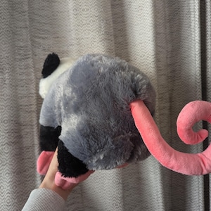 May include: A plush gray and white opossum toy with a pink curled tail. The toy has a black nose and black eyes.