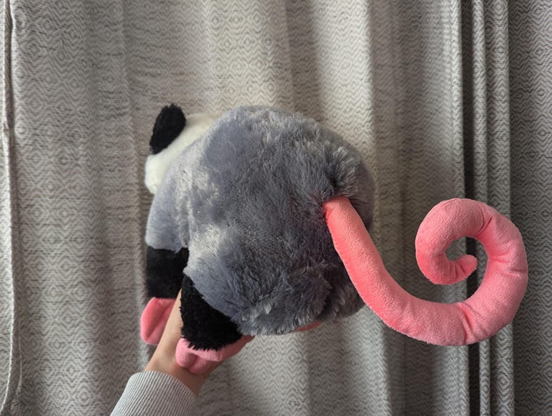 May include: A plush gray and white opossum toy with a pink curled tail. The toy has a black nose and black eyes.