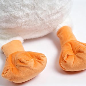 May include: A plush toy featuring a soft white textured body and two bright orange webbed feet. The stuffed animal has a rounded, fluffy white base and smooth orange fabric on the feet, showcasing a simple design with distinct color contrast.