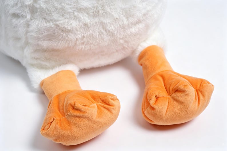 May include: A plush toy featuring a soft white textured body and two bright orange webbed feet. The stuffed animal has a rounded, fluffy white base and smooth orange fabric on the feet, showcasing a simple design with distinct color contrast.
