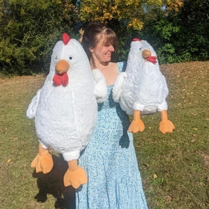 May include: Two white plush chicken toys. The larger chicken is being held by a person wearing a blue and white floral dress. The smaller chicken is being held by the person's other hand.