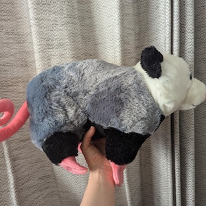 May include: A plush gray and white opossum toy with a pink curled tail. The toy is lying on its side with its head turned to the right.