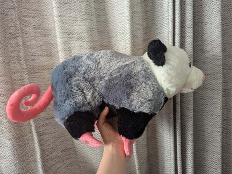 May include: A plush gray and white opossum toy with a pink curled tail. The toy is lying on its side with its head turned to the right.