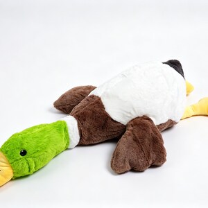 Puede incluir: Un juguete de peluche con forma de pato azul&oacute;n que presenta una cabeza verde brillante, un pico amarillo, un anillo blanco en el cuello y un cuerpo cubierto de suave pelaje marr&oacute;n y blanco. El peluche est&aacute; en posici&oacute;n tendida con alas.