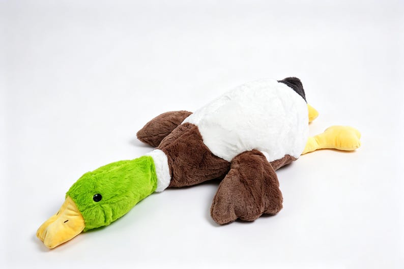 Puede incluir: Un juguete de peluche con forma de pato azul&oacute;n que presenta una cabeza verde brillante, un pico amarillo, un anillo blanco en el cuello y un cuerpo cubierto de suave pelaje marr&oacute;n y blanco. El peluche est&aacute; en posici&oacute;n tendida con alas.