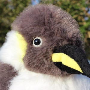 May include: A plush penguin toy featuring soft grey fur on the head and back, contrasted by white fur on the chest and neck. The face includes a black beak with a bright yellow patch, a round black eye with a white iris, and yellow accents.