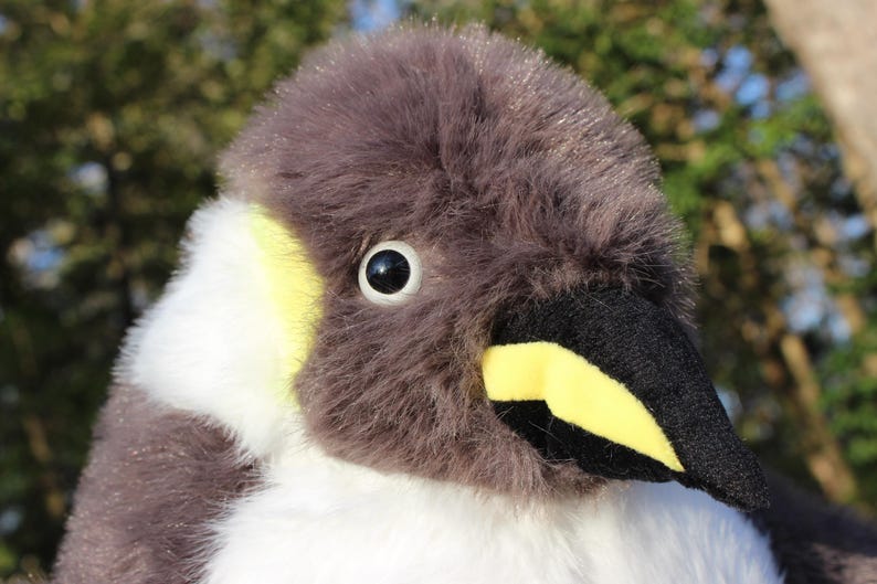 May include: A plush penguin toy featuring soft grey fur on the head and back, contrasted by white fur on the chest and neck. The face includes a black beak with a bright yellow patch, a round black eye with a white iris, and yellow accents.