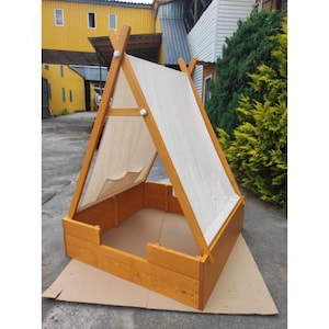 Op de afbeelding: Een houten zandbak met een tipi-stijl luifel. De zandbak is gemaakt van lichtbruin hout en heeft een rechthoekige basis. De luifel is gemaakt van een lichtgekleurde stof en biedt schaduw. De structuur is ontworpen voor buitenspelen.