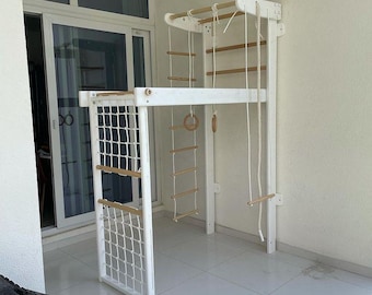 May include: White wooden indoor play structure with a climbing wall, rope ladder, and a net. The structure has a platform with a rope swing and a wooden ring.