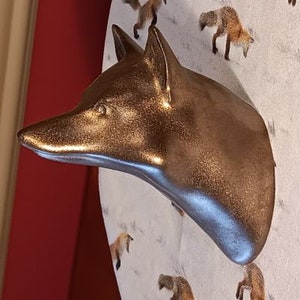 May include: A bronze-colored fox head sculpture with a metallic finish. The sculpture is mounted on a circular background with illustrations of foxes. The fox head has detailed features and is a decorative item.