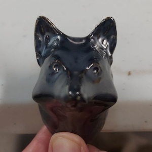May include: A ceramic fox head sculpture with a dark blue glaze. The sculpture has a realistic appearance with detailed features, including the eyes, nose, and ears.