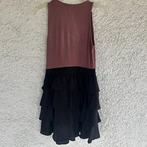 Puede incluir: Vestido sin mangas con un corpi&ntilde;o malva y una falda negra con volantes en capas. El vestido est&aacute; colgado de una percha de pl&aacute;stico transparente sobre un fondo texturizado blanquecino. El vestido parece estar hecho de tela suave.